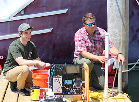 Two men sit on a dock with scientific equipment to collect nutrient data.
